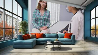 Lovely senior patient seated on medical chair while explaining to young latina doctor her health concern. Attractive mid aged woman consulting with young doctor about health condition Wall mural