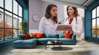 Lovely young hispanic woman looking at test results on tablet computer with senior doctor. Mid aged medical doctor using portable tablet to review health history of patient Wall mural