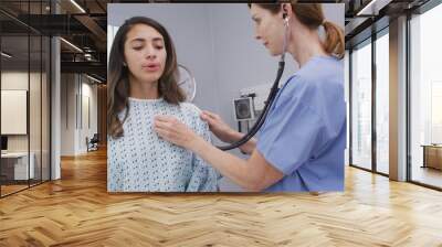 Mid aged nurse using stethoscope to examine young patients lungs and heart. Young latina patient takes deep breathes while nurse uses medical instrument to  check body vitals Wall mural