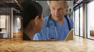 Over the shoulder shot of doctor with tablet computer explaining treatment to patient in hospital exam room. Senior Caucasian male medical professional and Asian woman Wall mural