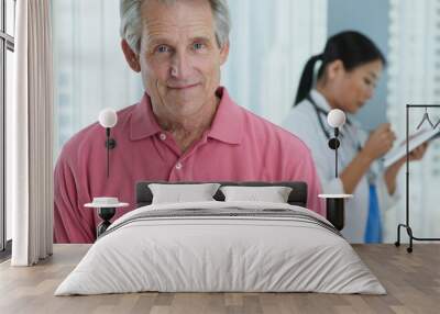 Portrait of senior Caucasian male patient looking at camera and smiling while doctor works in background. Attractive older man in hospital with female physician out of focus behind him Wall mural