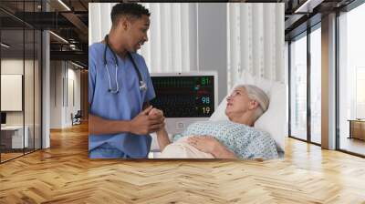 Portrait of young male nurse showing support of elderly senior patient in bed holding her hand. Registered nurse holding hand of ill female woman in hospice Wall mural