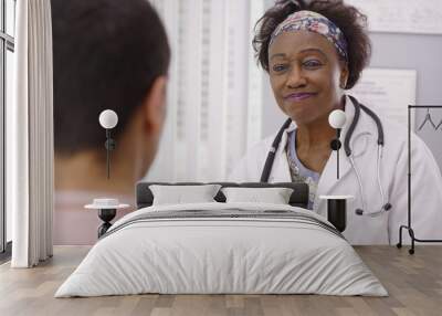 Positive female African doctor talking to male patient inside medical office. Close up of senior black MD smiling with young adult male patient and talking about health concerns Wall mural