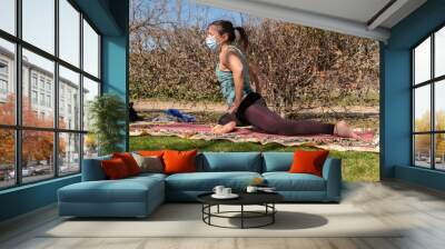 A brunette woman doing yoga class in a park on a sunny day and wearing a face mask Wall mural