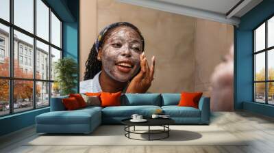 A young African woman smiles as she applies a face mask in a bathroom, enjoying a moment of self-care. Wall mural