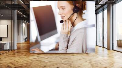 Businesswoman talking by headset in sunny office. Call center and diverse people group in business Wall mural