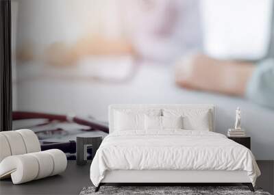 Doctor and patient are sitting and discussing something at the desk in the clinic office. The focus is on the stethoscope lying on the table, close up. Perfect medical service and medicine concept. Wall mural