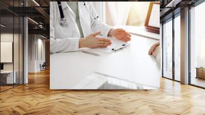 Doctor and patient sitting and discussing something near each other at the white desk in clinic. Female physician is listening filling up a records form. Medicine concept Wall mural