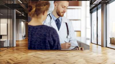 Friendly bearded doctor and patient woman discussing current health examination while sitting in sunny clinic. Medicine concept Wall mural