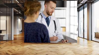 Friendly red-bearded doctor and patient woman discussing current health examination while sitting in sunny clinic. Medicine concept Wall mural