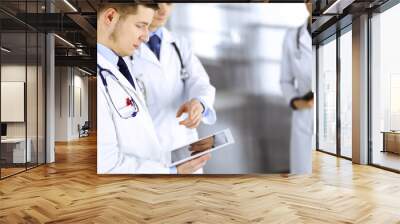 Group of doctors are checking medical names on a computer tablet, with a nurse with a clipboard on the background, standing together in a hospital office. Physicians ready to examine and help patients Wall mural