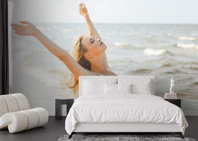 Happy blonde beautiful woman on the ocean beach standing in a white summer dress, raising hands Wall mural