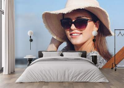 Happy brunette woman wearing sunglasses and hat relaxing on a wooden deck chair at the ocean beach Wall mural