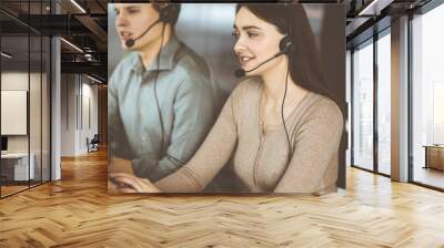 Two young people in headsets are talking to the clients, while sitting at the desk in an office. Focus on woman. Call center operators at work Wall mural