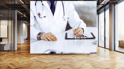 Unknown male doctor sitting and working with clipboard of medication history record in clinic at his working place, close-up. Young physician at work. Perfect medical service, medicine concept Wall mural