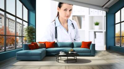 Young brunette female doctor sitting  with clipboard near window in  hospital and filling up medical history form. Wall mural