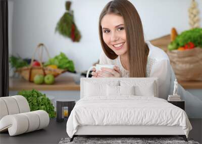 Young happy woman holding white cup and looking at the camera while sitting at wooden table in the kitchen among green vegetables. Good morning, lifestyle or cooking concept Wall mural