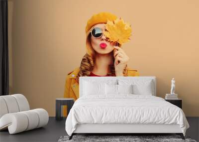 Autumn color style outfit, portrait of stylish beautiful young woman model with yellow maple leaves blowing her lips sends sweet kiss wearing orange french beret, sunglasses on brown studio background Wall mural