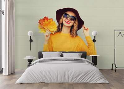 Autumn portrait of happy smiling young woman holds yellow maple leaves wearing round hat, sweater on gray background Wall mural