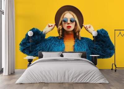 Fashionable portrait of stylish young woman, female model posing wearing blue faux fur, round hat on yellow background Wall mural