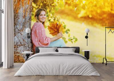 Happy smiling woman with yellow maple leafs sitting under tree i Wall mural