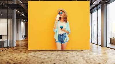 Happy smiling young woman 20s drinking coffee with smartphone looking away wearing summer straw hat, shorts on orange background Wall mural