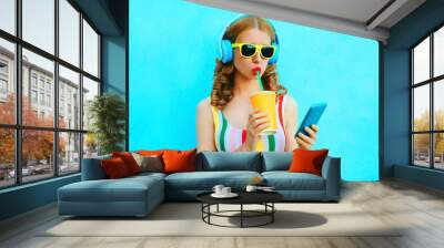 Portrait cool girl drinking fruit juice holding phone listening to music in wireless headphones on colorful blue background Wall mural