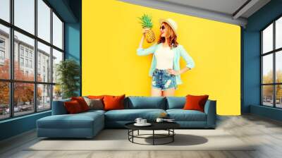 Portrait happy smiling woman holding pineapple having fun in summer straw hat, sunglasses, shorts on colorful yellow background Wall mural