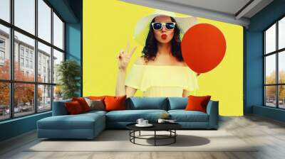 Portrait of beautiful young woman blowing her lips sending air kiss with red balloon wearing summer straw white hat on yellow background Wall mural