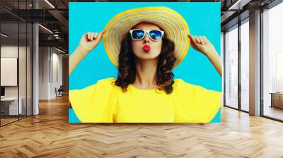 Portrait of beautiful young woman blowing her lips sending sweet air kiss posing wearing yellow summer straw hat, dress on blue background Wall mural