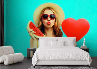 Portrait of beautiful young woman with slice of fresh watermelon and big red heart shaped balloon blowing her lips wearing summer straw hat, sunglasses on blue background Wall mural