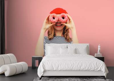 Portrait of happy funny smiling woman covering her eyes looking for something looking through donuts as binoculars having fun on pink background Wall mural