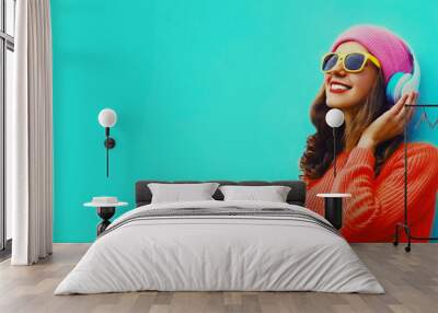 Portrait of happy smiling modern young woman in wireless headphones listening to music with smartphone wearing red knitted sweater, pink hat on blue background Wall mural