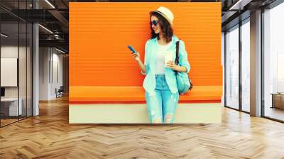 Portrait of happy smiling young woman with smartphone and cup of coffee wearing summer straw hat, backpack on orange background, blank copy space for advertising text Wall mural