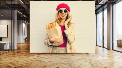 Portrait of happy surprised smiling woman holding grocery shopping paper bag with long white bread baguette wearing beret on blank gray background Wall mural