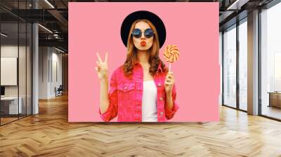 Portrait of happy young woman holding lollipop and blowing her lips sends air kiss wearing pink jacket, black round hat on pink background Wall mural