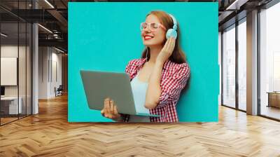 Portrait of modern young woman working with laptop listening to music in headphones on blue background Wall mural