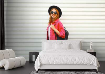 Portrait of stylish modern smiling young woman wearing pink jacket, black round hat and backpack on white background, blank copy space for advertising text Wall mural