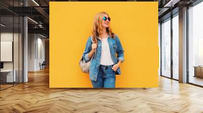 Portrait of stylish modern smiling young woman with backpack in denim jacket on yellow background Wall mural