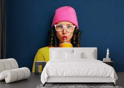 Portrait of stylish modern young woman drinking fresh juice wearing colorful clothes, yellow knitted sweater and pink hat on dark blue background, blank copy space for advertising text Wall mural