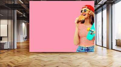 Portrait of stylish young woman with burger fast food and skateboard wearing baseball cap, sunglasses on pink background Wall mural