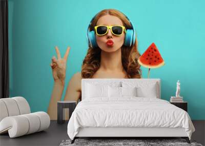 Summer fashion portrait of young woman in headphones listening to music with juicy slice of watermelon, female model blowing her lips posing on a colorful blue background Wall mural