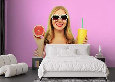 Summer portrait of happy smiling young woman drinking fresh juice with slice of juicy grapefruit wearing heart shaped sunglasses on pink background Wall mural