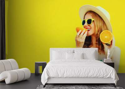 Summer portrait of young woman eating fresh slices of orange fruits wearing straw hat, sunglasses on yellow background, blank copy space for advertising text Wall mural