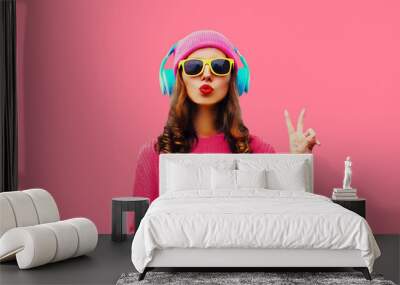 Trendy colorful portrait of stylish young woman in headphones listening to music with phone and blowing her lips sends air kiss on pink background Wall mural