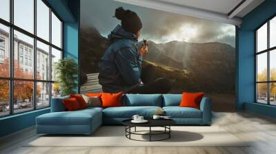 A hiker girl sits on the viewpoint and eats an energy bar, watching the sunset on the mountains Wall mural