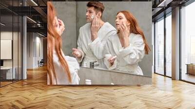 adorable caucasian couple take care of facial skin, woman applying cream while man is going to shave stubble Wall mural