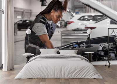 Afro Car mechanic woman is examining under hood of car at repair garage, wearing overalls, looking confident and concentrated. Side view on female trying to solve the problem of inoperative engine Wall mural