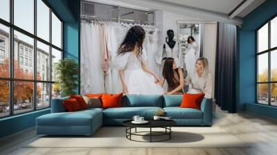 Beautiful bride is trying on an elegant wedding dress in modern wedding salon, her friends are helping Wall mural