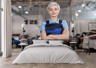 Caucasian professional female mechanic posing at camera standing in auto repair shop. Short haired woman in blue uniform. Car service, repair, maintenance and people concept. Front view. Copy space Wall mural